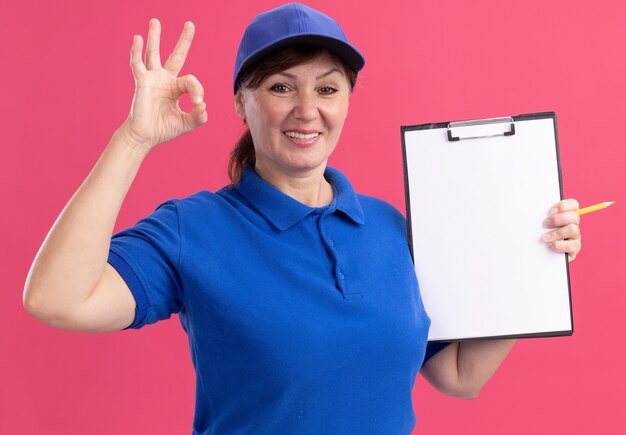 Middle aged delivery woman in blue uniform and cap holding clipboard with blank pages looking at front smiling cheerfully showing ok sign standing over pink wall