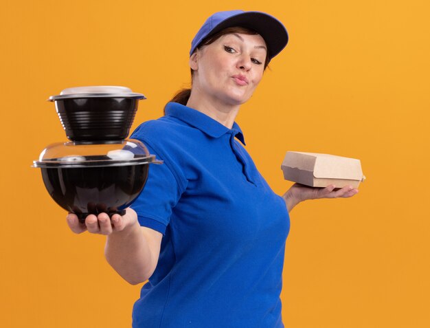 オレンジ色の壁の上に立って自信を持って笑顔に見える食品パッケージを保持している青い制服と帽子の中年配達の女性