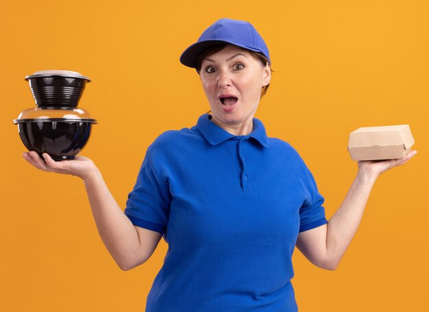 Middle aged delivery woman in blue uniform and cap holding food packages looking at front amazed and surprised standing over orange wall