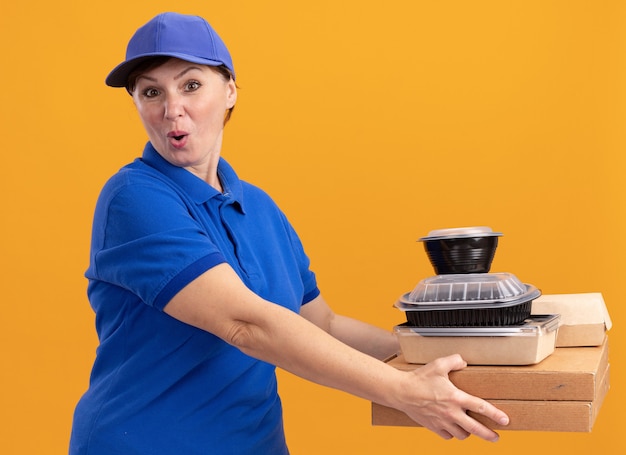 Женщина-доставщик средних лет в синей униформе и кепке держит коробки для пиццы и продуктовые пакеты, глядя вперед, счастливая и удивленная, стоя над оранжевой стеной