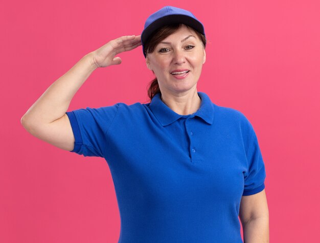 ピンクの壁の上に立って自信を持って敬礼を笑顔で正面を見て青い制服とキャップの中年配達の女性