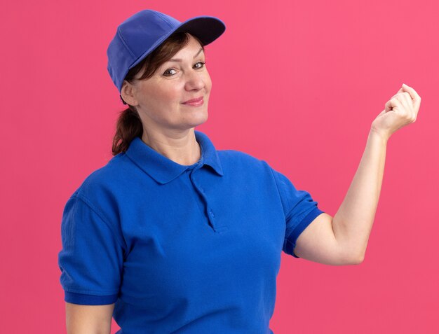 Middle aged delivery woman in blue uniform and cap looking at front smiling making money gesture rubbing fingers waiting for payment standing over pink wall