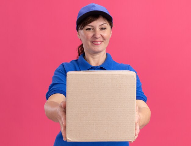 Middle aged delivery woman in blue uniform and cap showing cardboard box looking at front smiling confident standing over pink wall