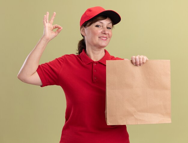 Middle aged delivery woman in red uniform and cap holding paper package looking at front smiling cheerfully showing ok sign standing over green wall