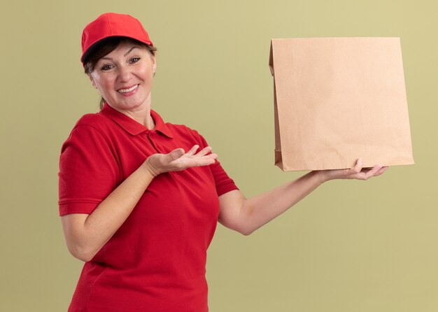 Middle aged delivery woman in red uniform and cap holding paper package presenting with arm looking at front smiling confident standing over green wall