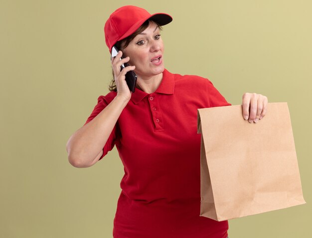Женщина-доставщик средних лет в красной форме и кепке держит бумажный пакет, разговаривает по мобильному телефону и выглядит смущенной, стоя у зеленой стены