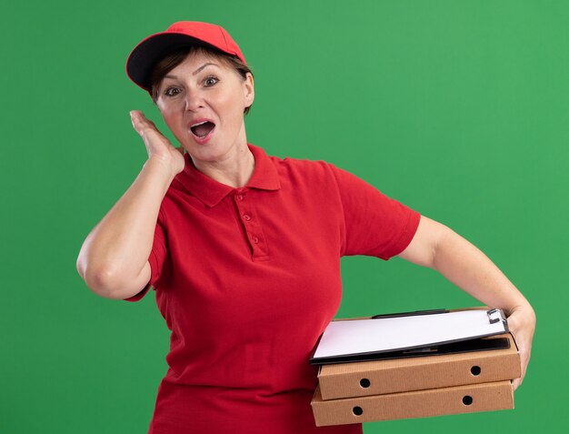 赤い制服を着た中年の配達の女性とピザの箱とクリップボードを保持し、空白のページが正面を見て混乱し、緑の壁の上に立って驚いた