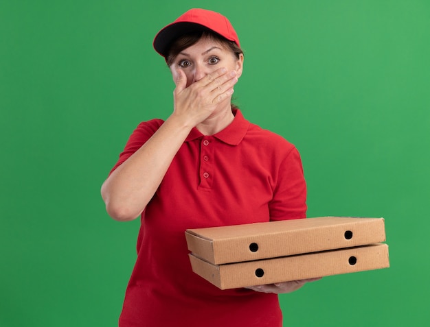 Middle aged delivery woman in red uniform and cap holding pizza boxes looking at front being shocked covering mouth with hand standing over green wall