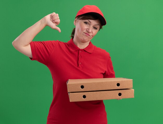 Middle aged delivery woman in red uniform and cap holding pizza boxes looking at front smiling pointing at herself standing over green wall