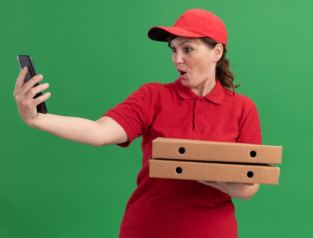 Женщина-доставщик средних лет в красной униформе и кепке держит коробки с пиццей и смотрит на смартфон, удивившись и смущаясь, стоя у зеленой стены