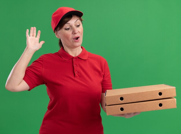 Middle aged delivery woman in red uniform and cap holding pizza boxes looking at them happy and excited with arm raised standing over green wall