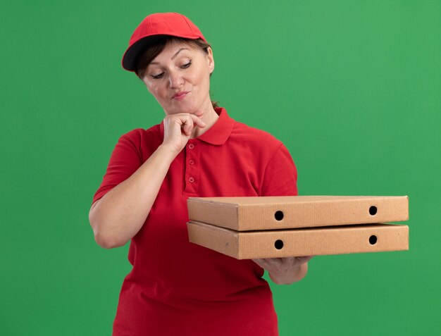 Middle aged delivery woman in red uniform and cap holding pizza boxes looking at them with pensive expression thinking standing over green wall