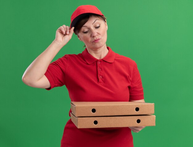 Middle aged delivery woman in red uniform and cap holding pizza boxes looking at them with serious face standing over green wall