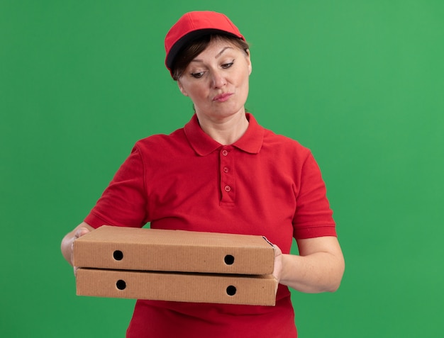 Женщина-доставщик средних лет в красной униформе и кепке держит коробки с пиццей и смотрит на них со скептическим выражением лица, стоя у зеленой стены
