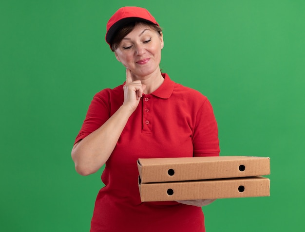 Женщина-доставщик средних лет в красной униформе и кепке держит коробки для пиццы, глядя на них с улыбкой на лице, счастливой и позитивной, стоя над зеленой стеной