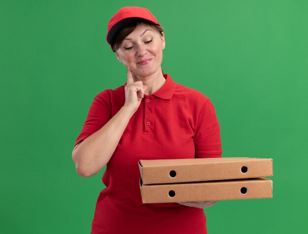 Middle aged delivery woman in red uniform and cap holding pizza boxes looking at them with smile on face happy and positive standing over green wall