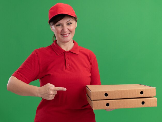 Middle aged delivery woman in red uniform and cap holding pizza boxes pointing with index finger at boxes smiling confident standing over green wall
