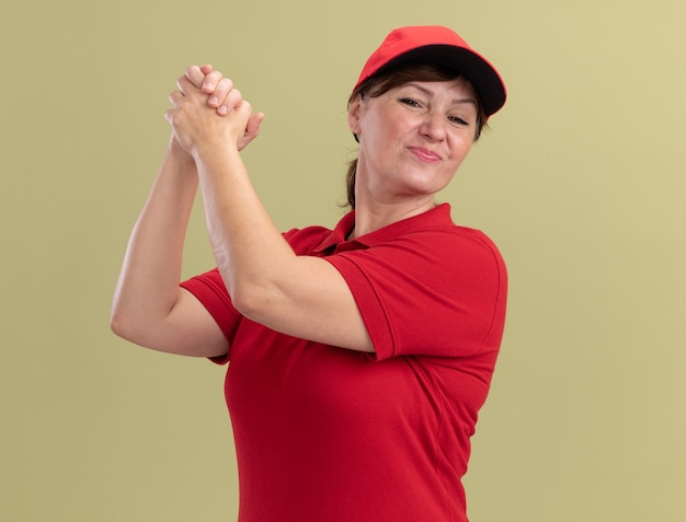 Middle aged delivery woman in red uniform and cap looking confident holding hands together making teamwork gesture standing over green wall