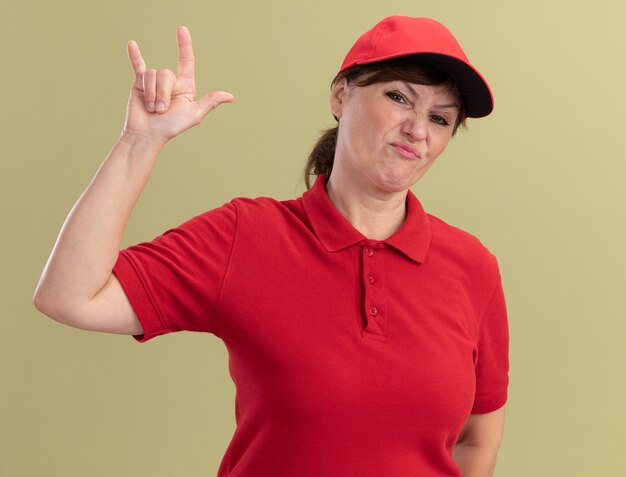 Женщина-доставщик средних лет в красной униформе и кепке, недовольно смотрящая на фронт, показывает символ скалы, стоящий над зеленой стеной
