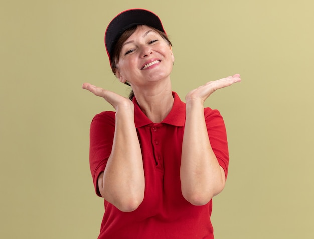 Middle aged delivery woman in red uniform and cap looking at front happy and positive smiling with hands raised standing over green wall