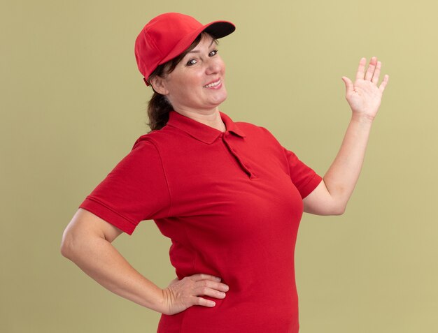 Женщина-доставщик средних лет в красной форме и кепке смотрит вперед, представляя пространство для копирования с рукой, стоящей над зеленой стеной