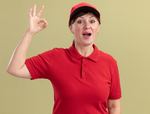 Middle aged delivery woman in red uniform and cap looking at front smiling cheerfully showing ok sign standing over green wall