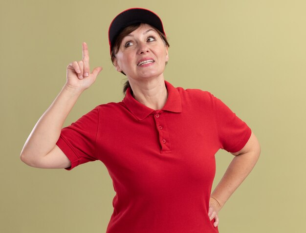 Женщина-доставщик средних лет в красной униформе и кепке, глядя вперед, уверенно улыбается, показывая указательный палец, имеющий новую отличную идею, стоящий над зеленой стеной