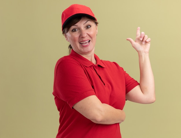 Middle aged delivery woman in red uniform and cap looking at front smiling confident showing index finger having new great idea standing over green wall
