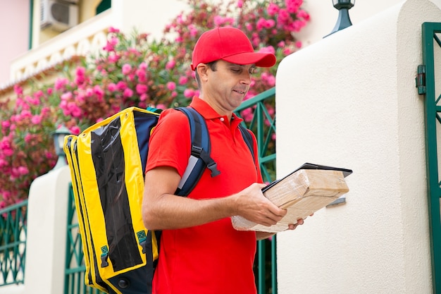 Middle-aged deliveryman holding clipboard and carton box and reading address in receipt. Focused postman in red uniform carrying thermo bag and delivering order. Home delivery service and post concept