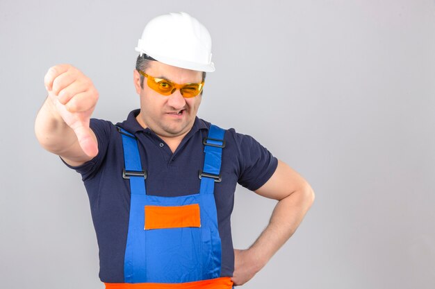 Middle aged displeased builder man wearing construction uniform and safety helmet showing thumbs down over isolated white wall