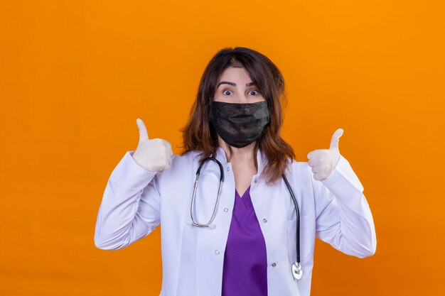 Доктор средних лет, одетый в белое пальто в черной защитной маске для лица и со стетоскопом, счастлив и показывает палец вверх обеими руками над оранжевым фоном
