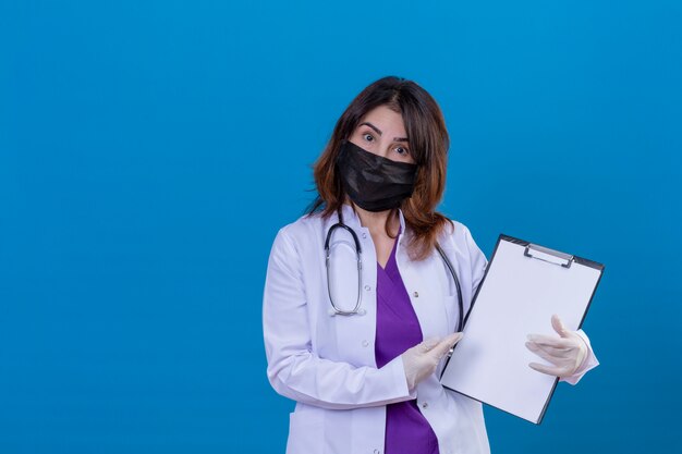 Middle aged doctor wearing white coat in black protective facial mask and with stethoscope holding clipboard pointing with index finger