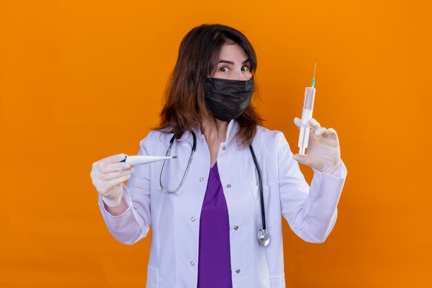Medico invecchiato centrale indossando camice bianco in maschera protettiva nera e con stetoscopio tenendo termometro digitale