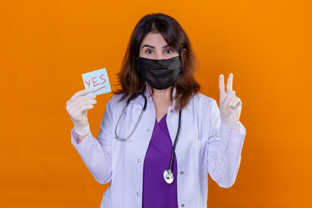 Middle aged doctor wearing white coat in black protective facial mask and with stethoscope holding reminder paper with yes word