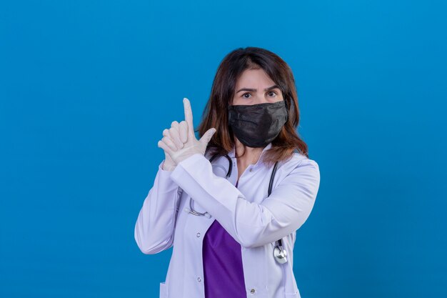 Middle aged doctor wearing white coat in black protective facial mask and with stethoscope holding symbolic gun with hand gesture