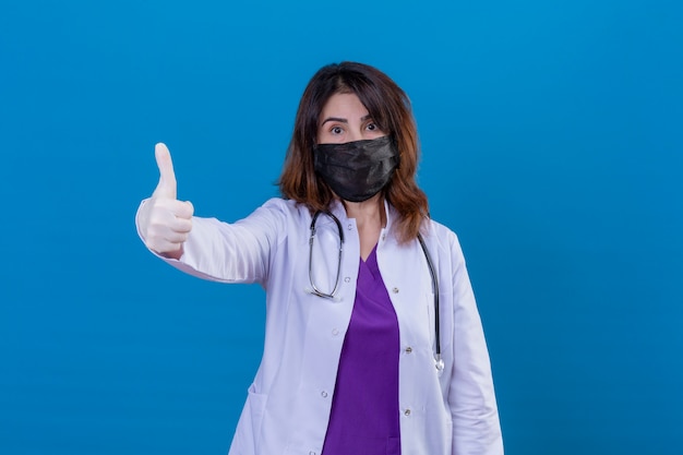 Middle aged doctor wearing white coat in black protective facial mask and with stethoscope looking positive showing thumbs up over blue wall