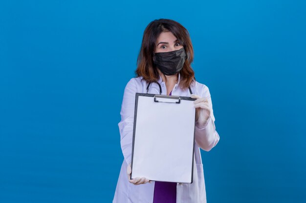Free photo middle aged doctor wearing white coat in black protective facial mask and with stethoscope showing clipboard