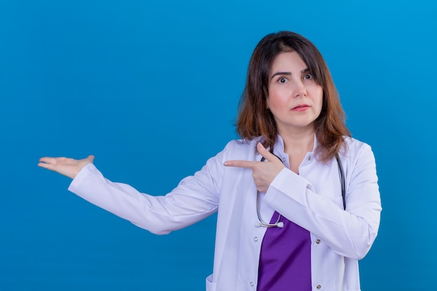 Middle aged doctor wearing white coat and with stethoscope looking confused pointing with both hands and finger to the side over blue wall
