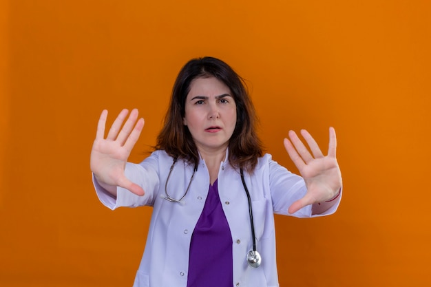 Доктор средних лет в белом халате и со стетоскопом с открытыми руками делает знак остановки с серьезным и уверенным выражением
