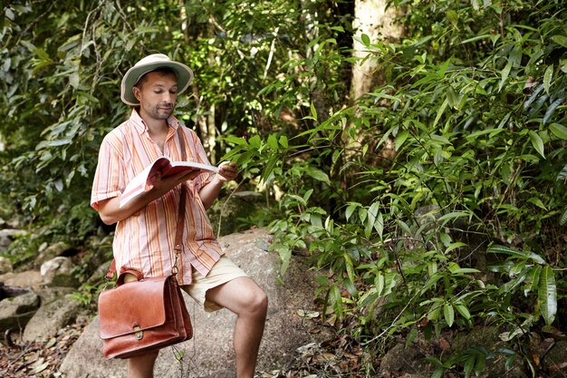 Middle aged European biologist or ecologist wearing hat and briefcase reading notes in his notebook during environmental studies outdoors, conducting research of plants, exploring tropical forest