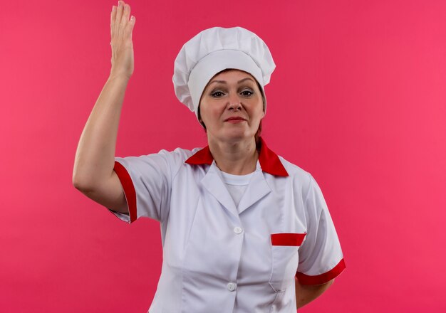  middle-aged female cook in chef uniform raising hand on isolated pink wall