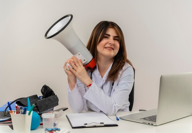 middle-aged female doctor wearing wearing medical robe with stethoscope sitting at desk work on laptop with medical tools holding loudspeaker on white wall with copy space