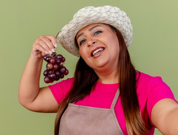 Middle aged gardener woman in apron and hat holding bunch of grape with smile