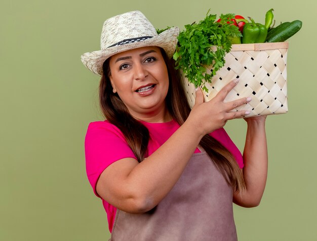 Middle aged gardener woman in apron and hat holding crate full of vegetables