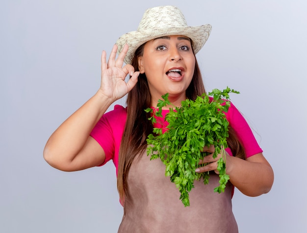 Giardiniere di mezza età donna in grembiule e cappello azienda erbe fresche guardando la telecamera sorridendo allegramente mostrando segno ok in piedi su sfondo bianco