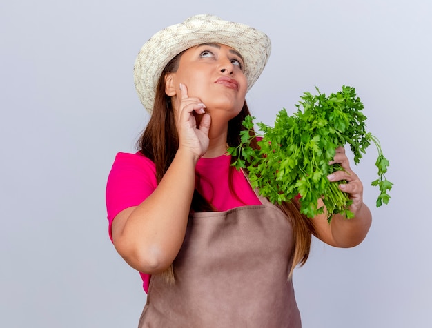 Middle aged gardener woman in apron and hat holding fresh herbs looking up puzzled standing over white background