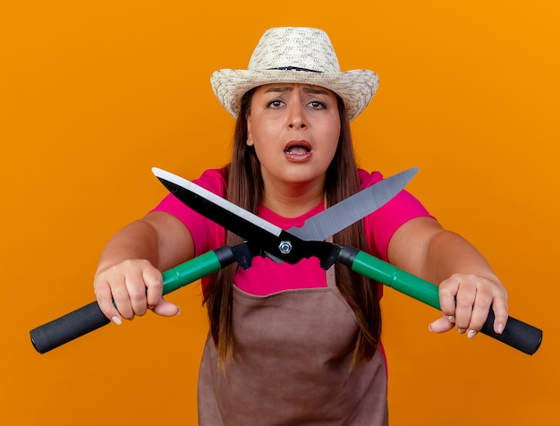 Middle aged gardener woman in apron and hat holding hedge clippers looking at camera being worried standing over orange background