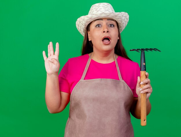 Middle aged gardener woman in apron and hat holding mini rake