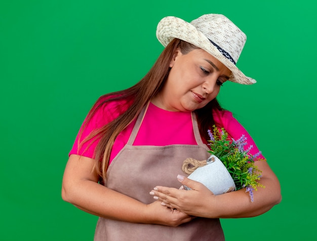 笑顔の赤ちゃんのように鉢植えの植物を保持しているエプロンと帽子の中年庭師の女性