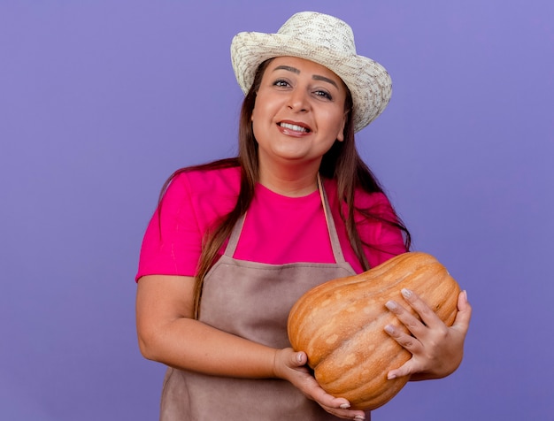Middle aged gardener woman in apron and hat holding pumpkin smiling cheerfully